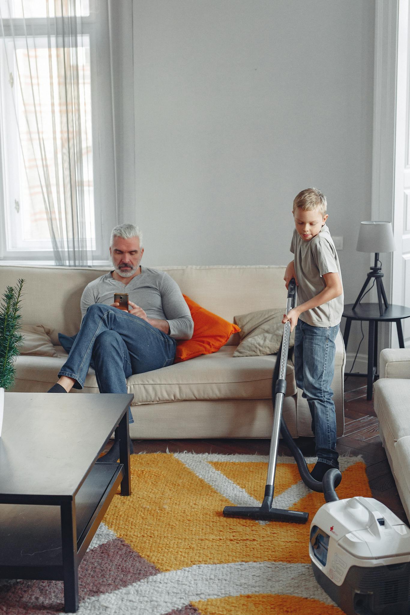 A cozy living room with father relaxing and son vacuuming, showing family bonding.