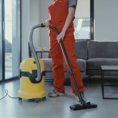A professional cleaner in red coveralls vacuuming a modern living room with a yellow vacuum cleaner.