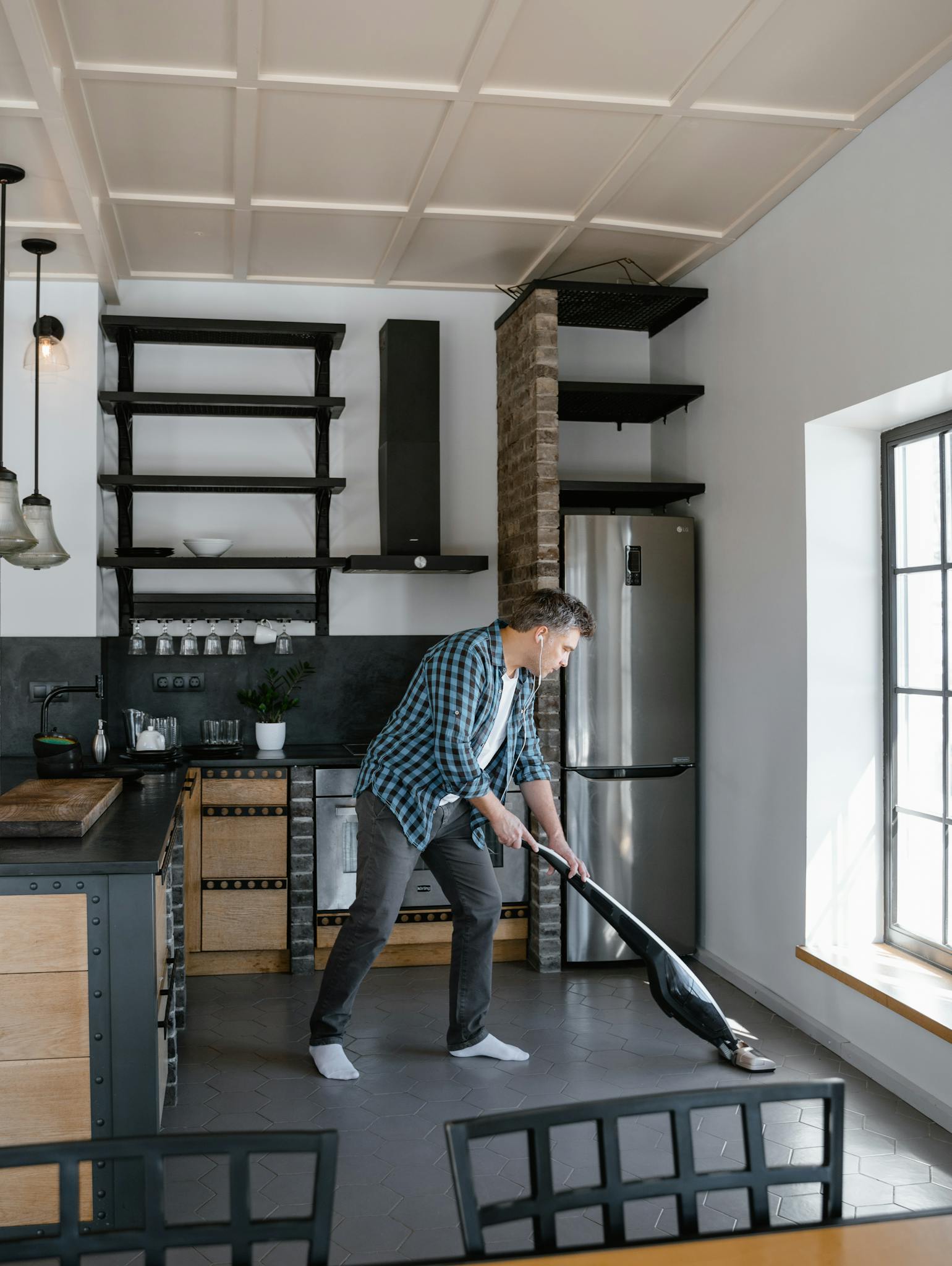 Adult man vacuuming a spacious, modern kitchen with natural light streaming in through large windows.