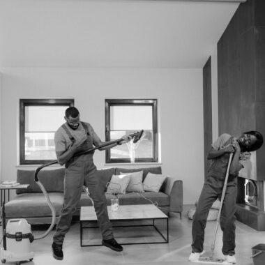 Two men clean a modern living room with vacuum and mop, enjoying their work.
